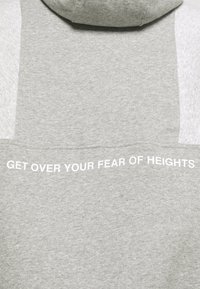 Ljusgrå hoodie med vit text på baksidan som lyder "ÖVERVINNA DIN RÄDSLA FÖR HÖJD" i versaler.