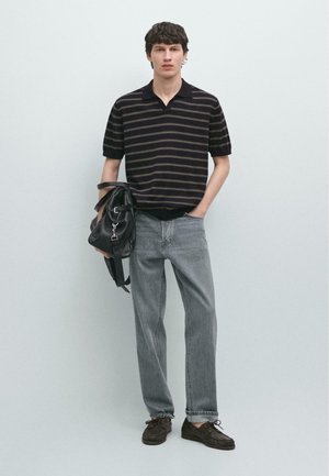 Giovane in piedi davanti a uno sfondo semplice, indossa una polo nera a righe, jeans grigi, tiene in una mano una borsa di pelle nera.