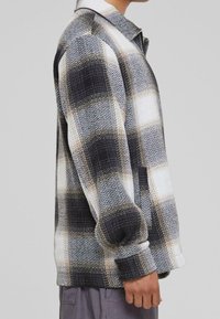 Koszulka w kratę z flaneli w kolorze czarnym, białym i szarym. Posiada kołnierzyk, długie rękawy oraz teksturowany wzór z subtelnymi akcentami w jodełkę.