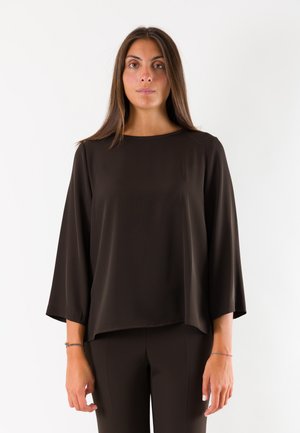 Donna con capelli castani lunghi che indossa una blusa nera larga a maniche lunghe e pantaloni neri, in piedi davanti a uno sfondo bianco uniforme.