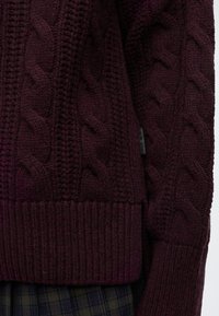 Manche et bord inférieur d'un pull torsadé violet foncé sur un fond en tissu à carreaux foncé.