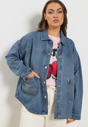 Vrouw die een oversized blauwe spijkerjack draagt over een roze t-shirt met afbeelding en crèmekleurige broek, staand tegen een effen lichte muur.