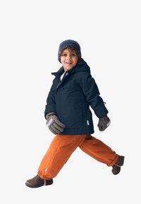 Blaue isolierte Jacke, orangefarbene wasserdichte Hose, graue Handschuhe, braune Winterstiefel und eine blaue Strickmütze. Kind in Gehpose.