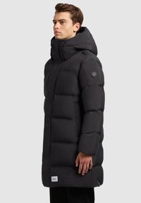 Abrigo puff negro con un cuello alto, delantero con cremallera y capucha ajustable. Presenta secciones acolchadas y un parche de logo en la manga.