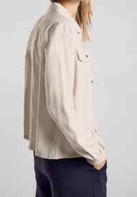 Kvinna iklädd en beige långärmad skjorta med knappslå och bröstfickor samt mörka byxor, stående med en hand i fickan mot en grå bakgrund.