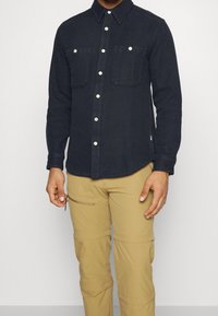 Mörk denimskjorta med långa ärmar, knapplist, två bröstfickor och kontrasterande knappar, ihopparad med ljusbeiga cargobyxor.