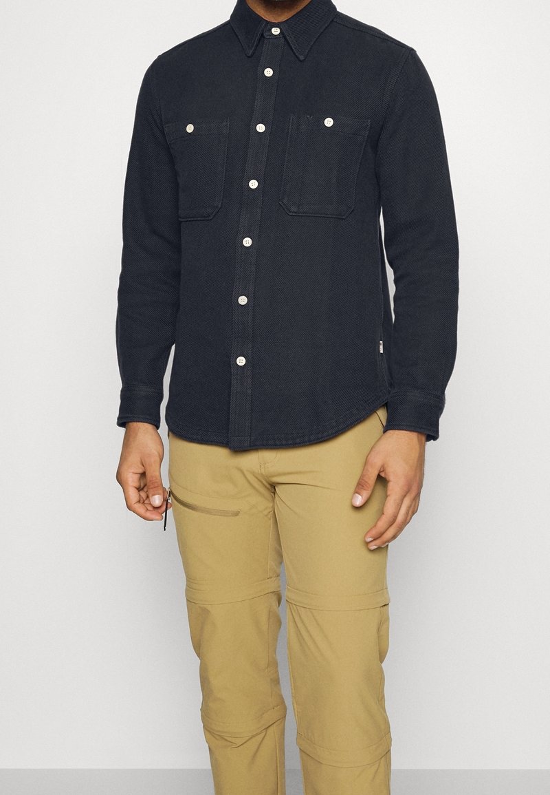 Mörk denimskjorta med långa ärmar, knapplist, två bröstfickor och kontrasterande knappar, ihopparad med ljusbeiga cargobyxor.