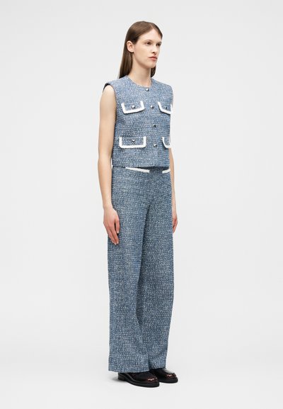 Νέα γυναίκα που φοράει μπλε γιλέκο από τουίντ χωρίς μανίκια με τσέπες με καπάκι και λευκή επένδυση, καθώς και παντελόνι με φαρδιά πόδια που ταιριάζει, στέκεται μπροστά σε απλό φόντο.