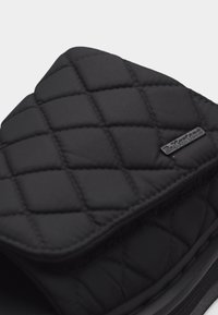 Μαύρο λουράκι σανδάλι με υφή από καπιτονέ ύφασμα και μικρή μαύρη ετικέτα με το λογότυπο "Dr. Martens" στο πάνω μέρος.