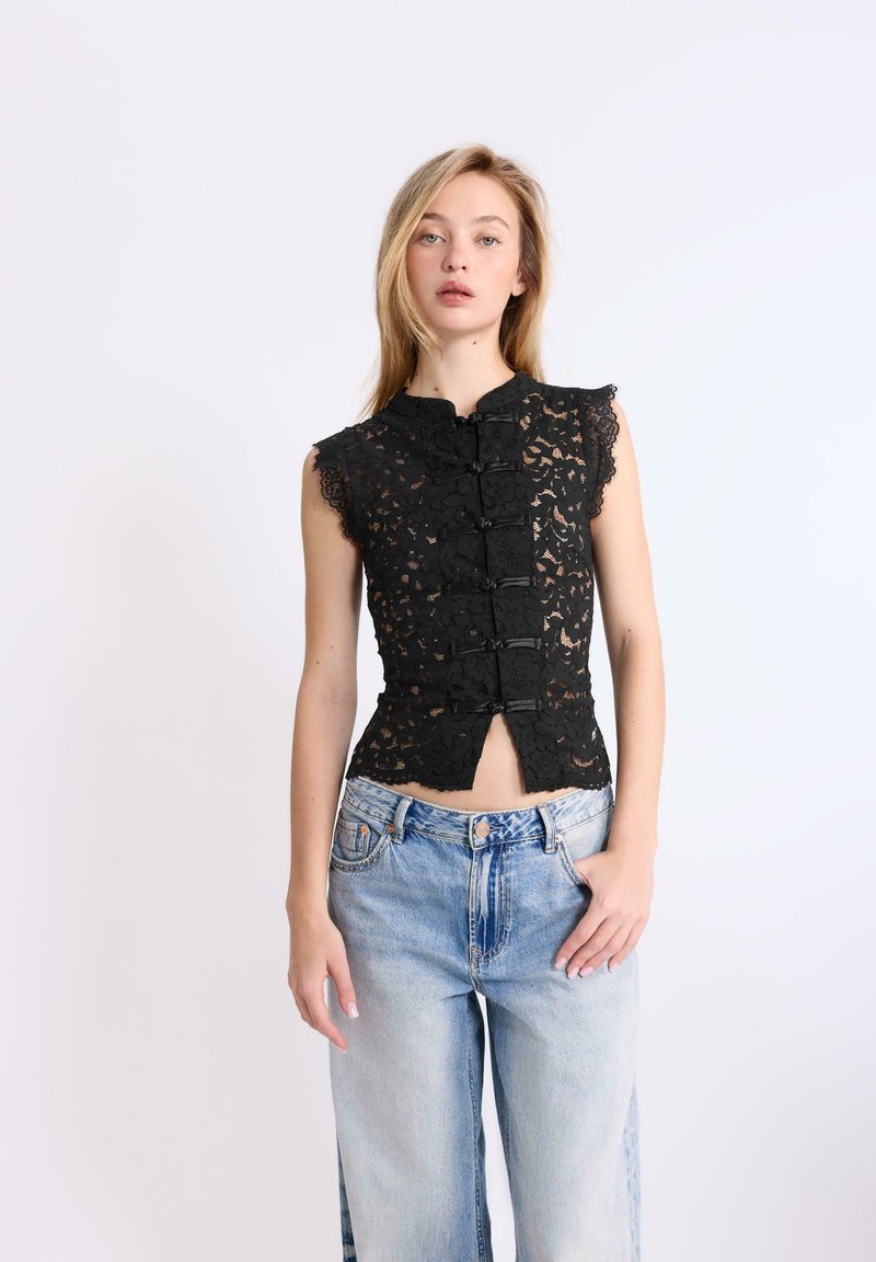Femme portant un haut en dentelle noire sans manches avec fermetures à boutons grenouille et un jean bleu clair taille haute, debout devant un fond uni.