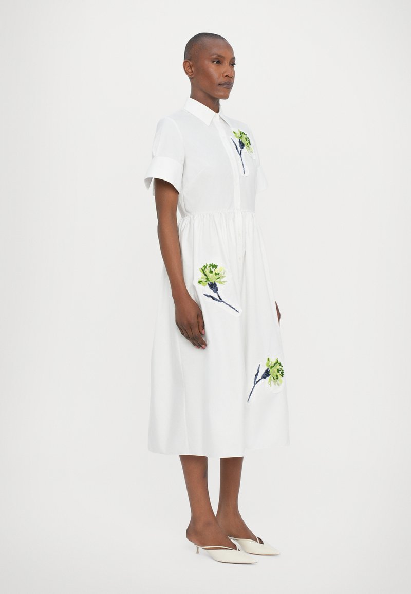 Abito di cotone bianco con maniche corte e chiusura a bottoni davanti. Presenta un ricamo floreale colorato sulla gonna. Abbinato a scarpe bianche.