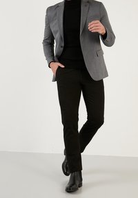 Šedý blazer s dvěma knoflíky přes černý rolák, spárovaný s černými slim-fit kalhotami a černými kotníkovými botami, stojící na neutrálním pozadí.