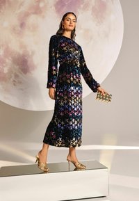 Rochie lungă, cu paiete, prezintă un model cu stele în nuanțe de albastru, mov și aur. Asortată cu pantofi cu toc înalt aurii și un clutch metalic.