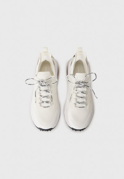 White mesh sneakers with textured accents, black rubber sole, and patterned laces. Features a pull tab at the heel for easy wear.