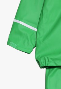Casaco de chuva verde com uma textura lisa e impermeável, apresentando uma faixa refletora branca na manga e punhos elásticos para um ajuste confortável.