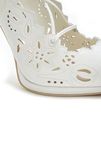 White high-heeled shoe made of leather, featuring cut-out floral patterns and scalloped edges. Decorated with gold accents and lace-up detail.