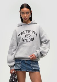 Felpa grigia con cappuccio, caratterizzata da una tasca a marsupio frontale, decorata con la scritta "WRSTBHV STUDIO" in nero, abbinata a una minigonna di jeans con tasche.