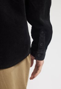 Zwarte corduroy shirt met verticale ribbels, knoopmanchetten en een ronde zoom. Gepaard met lichtbruine broek.