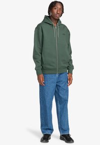 Grøn zip-up hoodie lavet af tykt stof, med hætte, snører og forlommer. Båret med løse blå denimjeans.