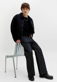 Ung person iført sort jakke, mørke vide jeans, sorte støvler og sølvkædehalskæde, siddende sidelæns på en metalstol.