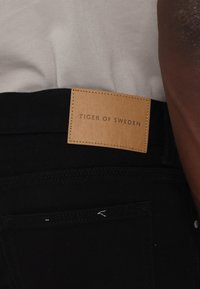 Čierne denimové rifľové nohavice s béžovou koženou nášivkou, na ktorej je embosovaný text "TIGER OF SWEDEN", s prešívaním a hladkým povrchom.