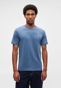 Zils īss piedurkņu kokvilnas T-krekls ar apaļu kakla izgriezumu un mazu baltu logotipu kreisajā krūšu daļā. Gluda tekstūra, ikdienišķa piegriezuma forma.