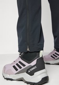 Pantalons légers noirs avec fermeture éclair à la cheville, associés à des chaussures de randonnée Terrex roses et grises avec des détails en maille et une semelle en caoutchouc.