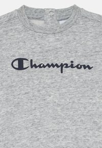 Svetilka v svetlo sivi barvi je izdelana iz teksturiranega materiala, s temno modrim logotipom "Champion" na sprednji strani in zapiranjem na gumbih pri vratu.