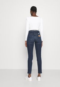 Skinny jeans in donkerblauwe denim met een hoge taille, achterzakken en een beige leren label op de tailleband, gecombineerd met witte sneakers.