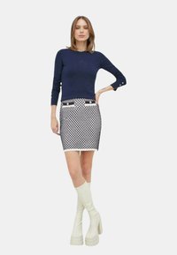 Maglione lavorato a maglia blu navy abbinato a una mini gonna bianca e blu con motivo, dotata di tasche frontali e bottoni; il modello indossa stivali bianchi fino al ginocchio.