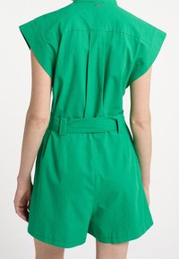 Combinaison verte sans manches avec taille ceinturée, plis au dos et détail col. Confectionnée en tissu léger, elle offre une coupe décontractée et un short.