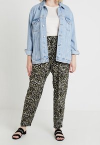 Lichtblauwe denimjack met twee borstzakken over een wit shirt, gecombineerd met loose-fit groene en zwarte broek met patroon en zwarte sandalen.