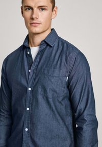 Dunkelblaues Chambray-Hemd aus strukturiertem Baumwollstoff. Mit Knopfleiste, weißen Knöpfen, langen Ärmeln und einer einzelnen Brusttasche.