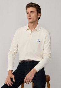 Polo a maniche lunghe crema con colletto a punta, caratterizzato da un logo blu ricamato. Indossato con jeans scuri e una cintura marrone. Tessuto liscio.
