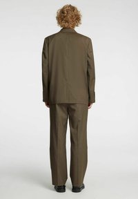 Brūns pielāgots uzvalks ar vienrindu žaketu un platām biksēm. Gluda tekstūra, aizmugurējā šķēluma detaļa un divu pogu aproces akcenti.