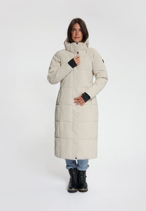 Vrouw draagt een lange beige gewatteerde winterjas met hoge kraag, blauwe spijkerbroek en zwarte veterlaarzen, staand tegen een witte achtergrond.