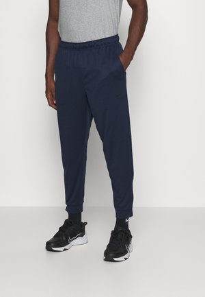 Homme portant un jogging bleu marine, des chaussures de sport noires, des chaussettes noires et un t-shirt gris, debout avec une main dans la poche.
