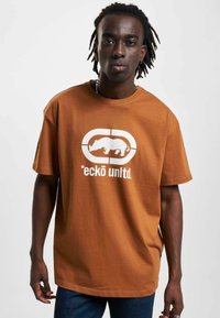 Brun bomulds T-shirt med en hvid grafik af en næsehorn og teksten "*eckō unlt.d." på forsiden. Korte ærmer, rund halsudskæring.
