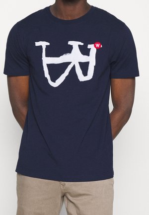 Mann trägt ein marineblaues T-Shirt mit einem großen weißen abstrakten Design und einem kleinen roten Kreis mit einem weißen "W" auf der Brust.