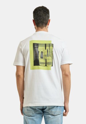 Uomo con capelli corti e ricci che indossa una maglietta bianca con un motivo astratto a quadrati gialli e neri sulla schiena, abbinata a jeans azzurri.