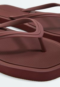 Tongs en caoutchouc marron texturé avec des brides épaisses et des entre-doigts arrondis, présentées en gros plan sur fond blanc.