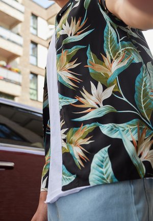 Person, die ein schwarzes Hemd mit großen tropischen Blumen- und Blattmustern trägt und neben einem roten Auto und einem Wohnhaus steht.