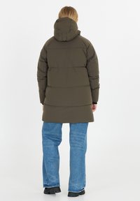 Olivgrön pufferjacka med huva, som har en quiltad design, axelpatchar och en medellång längd, bärs över blå vida jeans.