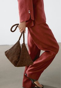 Osoba w luźnych czerwonych spodniach i bluzce, trzymająca brązową torebkę z koralików, idąca po gładkiej podłodze w czarnych sandałach.