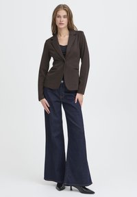 Blazer marrone con finitura testurizzata, chiusura con un solo bottone, rever a incrocio e due tasche frontali, abbinato a jeans scuri a gamba larga.