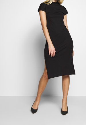 Femme portant une robe midi noire à manches courtes avec des fentes sur les côtés et des talons noirs à bout pointu, posant devant un fond blanc.