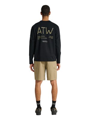 Mand stående med ryggen til iført sort langærmet skjorte med teksten "ATW", beige cargoshorts, sorte sokker og vandresko på hvid baggrund.