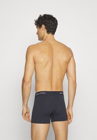 Musta puuvillainen bokserimalli, jossa on logolla varustettu vyötärö, tiukka istuvuus ja sileä pinta. Näkymä takaa, jossa näkyy vain vähän yksityiskohtia.