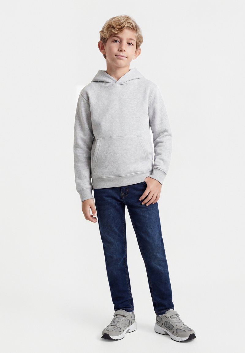 Băiat stând în picioare, purtând un hanorac gri deschis, blugi albastru închis și adidași gri, pe un fundal alb simplu.