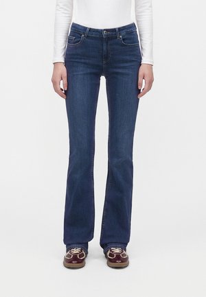 Person iført mørkeblå bootcut-jeans, hvid langærmet top og brune snørebåndssko, stående på en ensfarvet hvid baggrund.